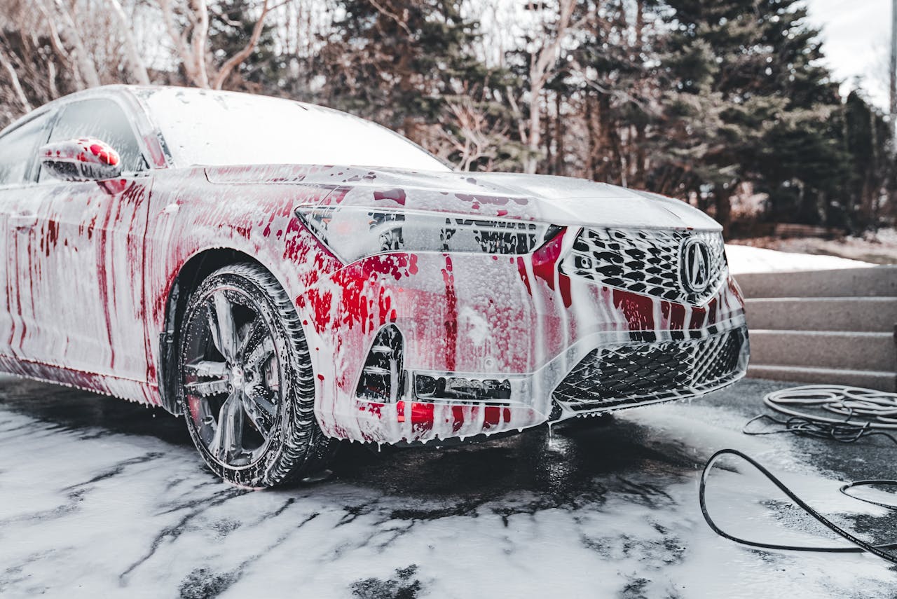 why-choose-us A vibrant red car covered in soapy foam during a thorough outdoor wash under daylight.