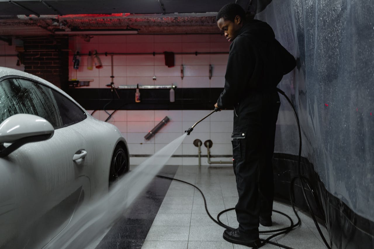 services-03 African American man cleaning a white car with a pressure hose in an indoor garage setting.